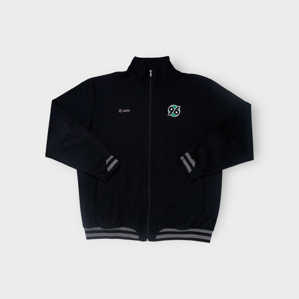 Black Jako track jacket with embroidered green Hannover 96 crest and grey sleeve stripes
