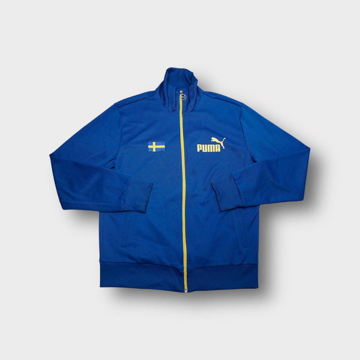 Blue Puma track jacket with yellow zipper, chest flag patch, and Puma logo