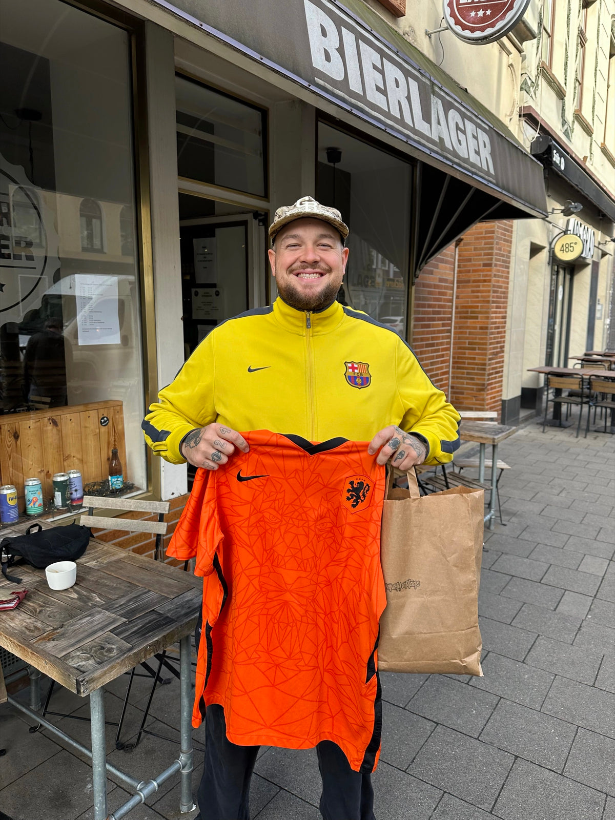 Lugatti in a yellow Barcelona track jacket, holding up an orange Netherlands jersey and a ninetyvintage bag.